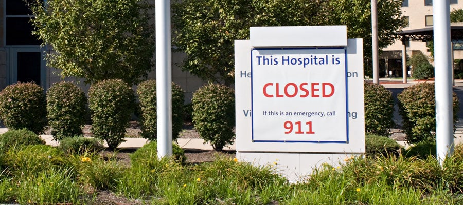 White sign covers the existing sign of in front of a hospital and says "This hospital is CLOSED, if this is an emergency call 911." The sign is flanked by shrubs and lamps posts.