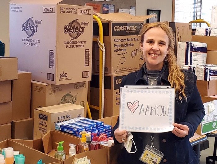 Female holding drawn sign with AAMOW on it infront of supplies for COVID-19