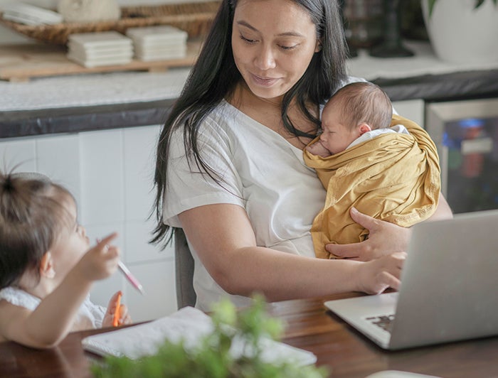 Digital Front Door. A mother sits at a kitchen counter while holding her baby and talking with her toddler. A laptop is open in front of the mother on which she is use a hospital's digital front door.