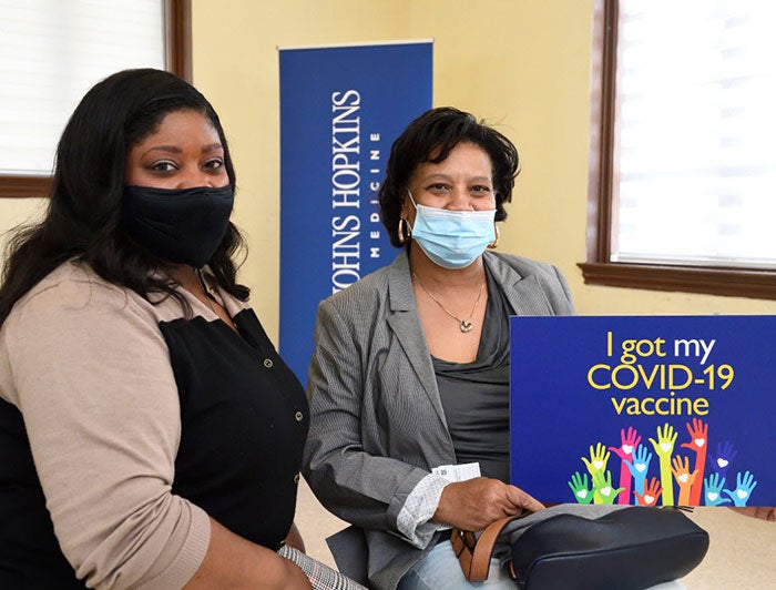 two women, wearing masks, sit at a Johns Hopkins vaccination event behind a sign that says 'I got my COVID-19 vaccine'