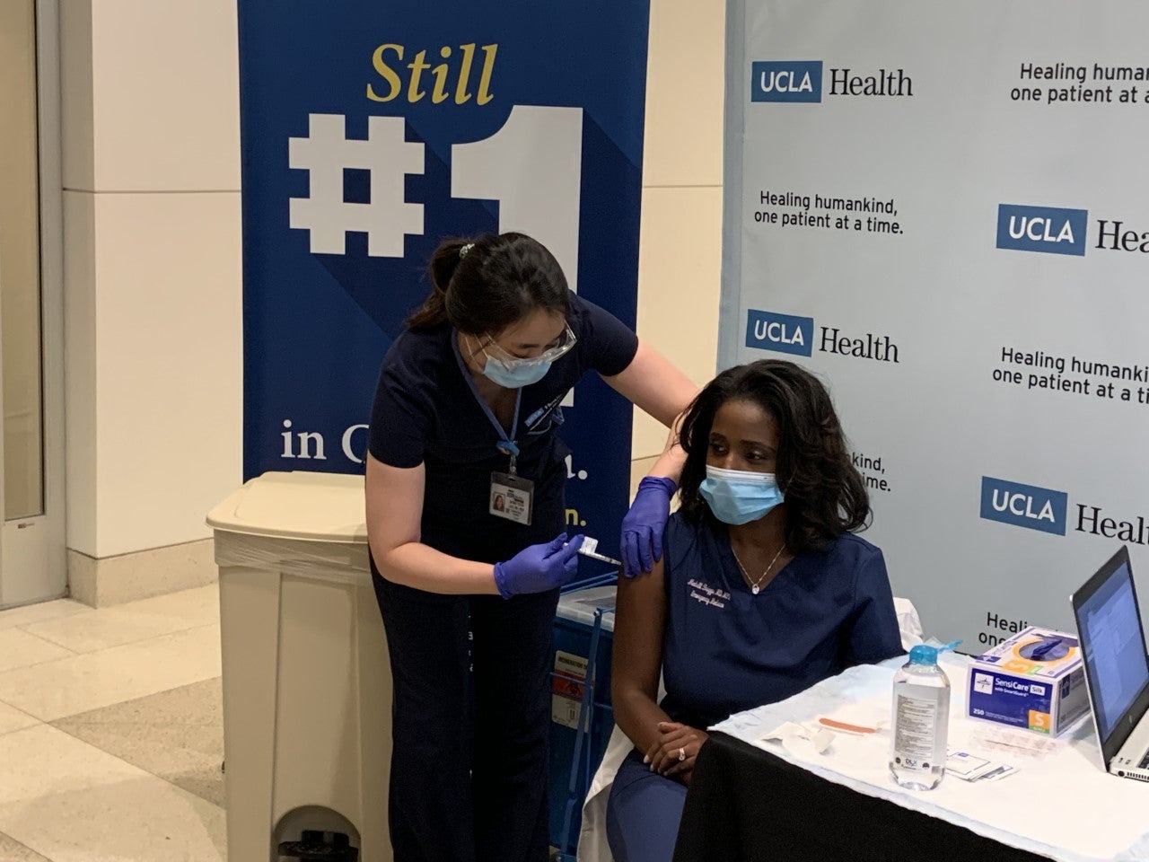 At a vaccination event, a female UCLA Health worker, wearing mask and scrubs, stands over a female colleague and adminsters COVID-19 vaccine