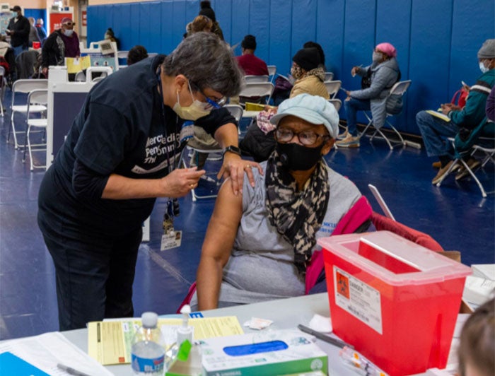 Penn Medicine health worker, wearing mask and scrubs, stands over a patient at a mass vaccnination event and adminsters COVID-19 vaccine