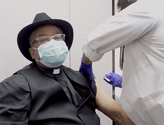 At a vaccination event, a female UCLA Health worker, wearing mask and scrubs, stands over a female colleague and adminsters COVID-19 vaccine