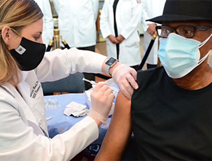 At a vaccination event, a female St. Dominic's health worker, wearing mask and scrubs, adminsters COVID-19 vaccine to a male patient wearing a fedora and medical mask