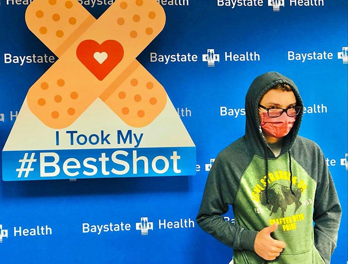Young patient in hoodie and mask is giving thumbs up while standing in front of Baystate Health Poster reading 'I took my #BestShot'