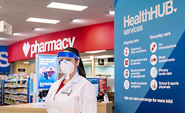 A Look Inside CVS Health’s Latest Moves to Disrupt Care. A clinician in a mask and face shield stands in front of a CVS pharmacy next to a HealthHUB Services sign.