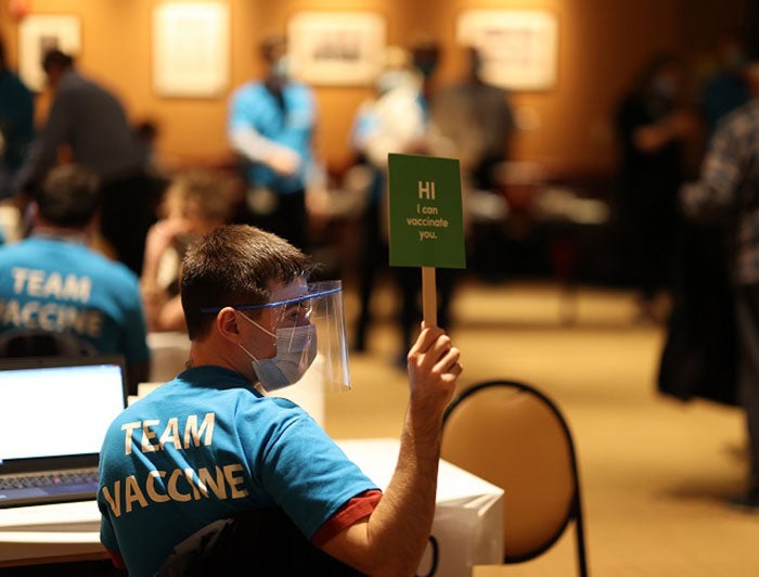 health worker wearing team vaccine shirt holds up sign that says they are available to give vaccine