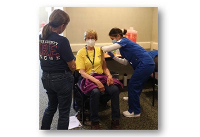 Female lifepoint patient sits in a chair while being vaccinated by health worker standing on right