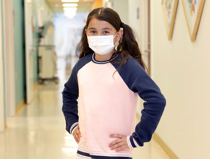 Young girl at Lurie, wearing a mask, stands with hands on hips like a superhero