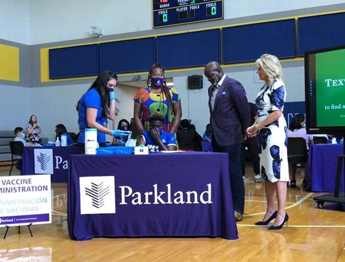 First Lady Jill Biden and Emmitt Smith stand in Emmett J. Conrad High School gymnasium next to table of students and Parkland staff