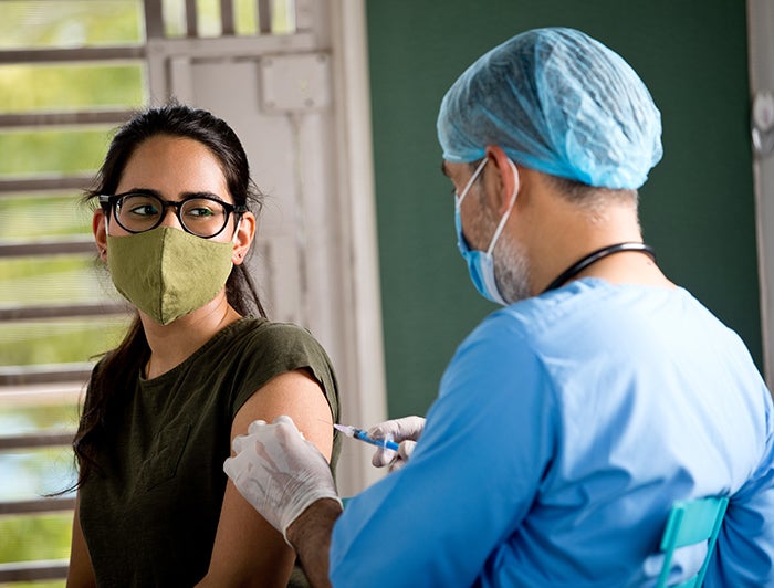 Female patient wearing glasses and medical mask is is adminstered shot by male health worker in scrubs and ppe