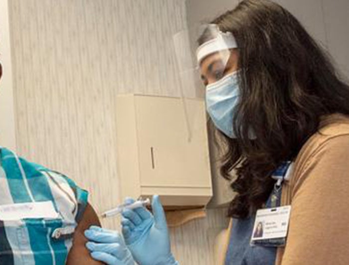 Female UR Medicine health worker wearing face shield, mask and gloves, gives jab in arm to patient