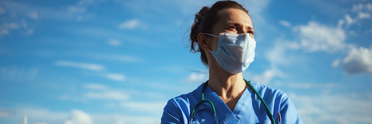 Emerging Lessons from the COVID-19 Pandemic. A medical clinician in scrubs and wearing a mask with a stethoscope wrapped around her neck stands with a blue sky behind her.