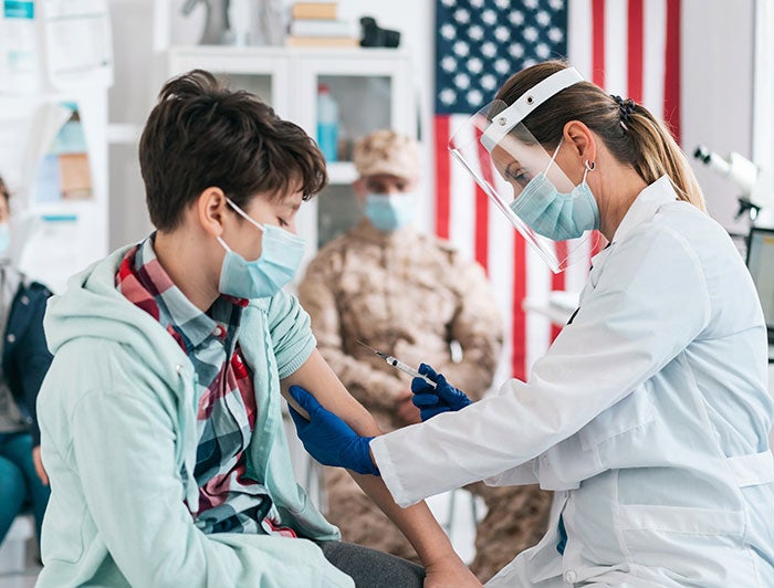 child being vaccinated by health worker in foreground, veteran family seated near American flag in clinic background