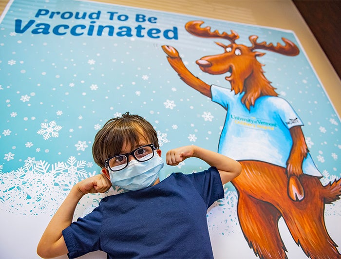 Young boy wearing medical mask stands with arms biceps flexed in muscle pose, beneath UVM poster that reads: Proud to be Vaccinated