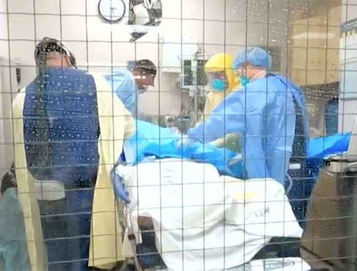 A team of health workers in PPE crowd around a patient, viewed through an inerior hospital room window