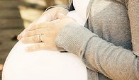 A pregnant woman stands with hands crossed on belly on a street lined with fallen leaves.