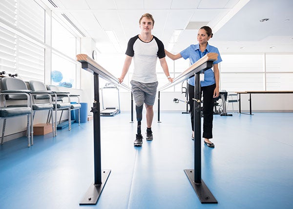 Post-Acute Rehabilitation. A man with a prosthetic leg walks between two support rails with the help of a physical therapist.