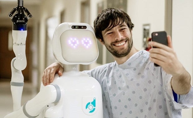 Collaborative Robot Aims to Ease Stress on Nurses. A patient takes a selfie on his mobile phone and Moxi, a health care robot.