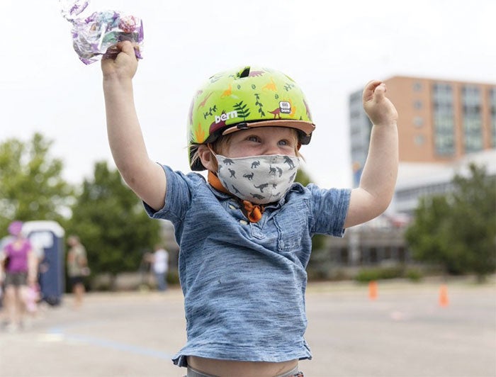 young white boy in mask and bike helmet raises arms in excitement