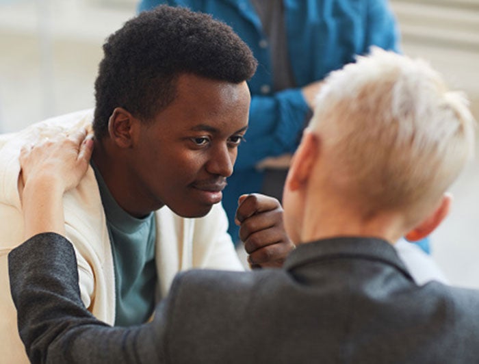 Young black man sits facing viewer, listening to person in suit jacket facing away from viewer.