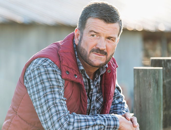 bearded man in flannel shirt and puffy vest stands leaning agains farm fence post