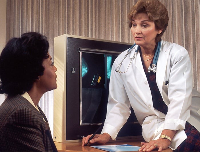 Woman in lab coat and stethoscope sits on the edge of a desk talking to seated woman