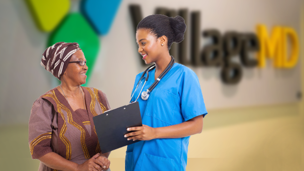 VillageMD Builds Bigger Footprint in the Northeast, Expands Value-Based Care Vision. A Black female patient wearing glasses and a Black female clinician with a stethoscope around her neck who is holding a chart stand in front of a wall with the VillageMD logo on it.