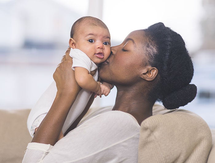 an african american mother, eyes closed, kisses her newborn baby on the cheek. The baby, eyes open, is looking toward viewer.