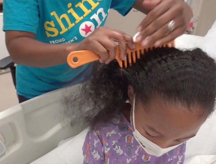 A Black adult woman is shown combing a black child's hair with a wide-toothed comb