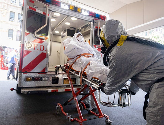 Health worker in full-body PPE loads an equally-protected patient into an ambulance