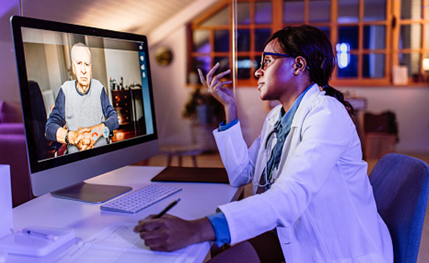 Virtual Multidisciplinary Rounds Cut Length of Stay, Costs. A doctor sitting at a desk and taking notes talks to a patient who appears on a computer monitor via virtual care telehealth technology.