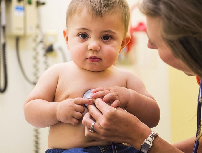 A round-cheeked baby sitting in a clinic office with a health worker holding stethoscope to baby's chest