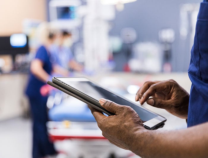 Stock photo of health worker in blue scrubs holding a table computer, nurses station in the background