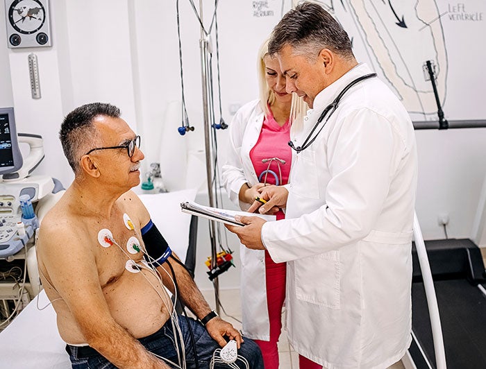 Cedars Sinai. Clinicians stand near patient bedside reviewing notes as patient sits wearing BP cuff and EKG leads