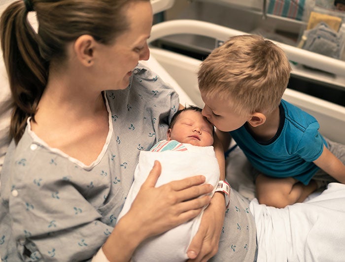 A woman in hospital gown sits in bed with newborn baby and a toddler kissing the baby's head