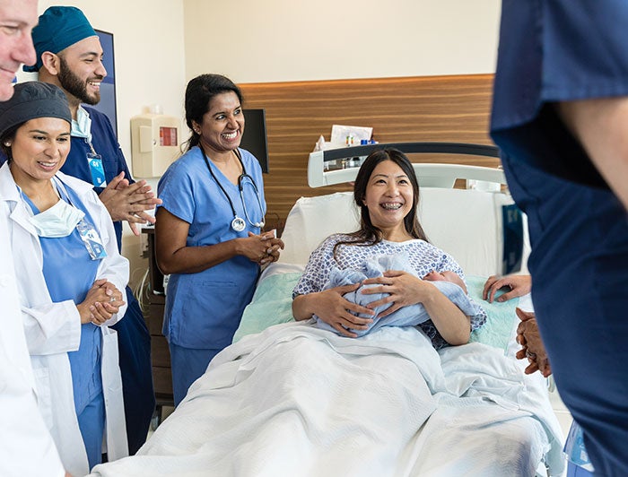 A mother holds baby in an L&D unit bed, surrounded by diverse medical staff