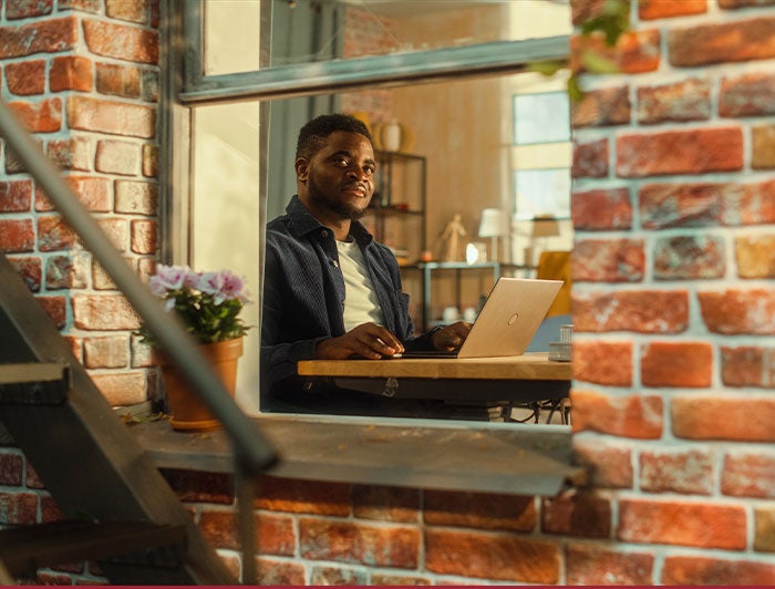 Kaiser Permanente. A young black man sitting in an apartment at a table working on a laptop, viewed through a fire escape window