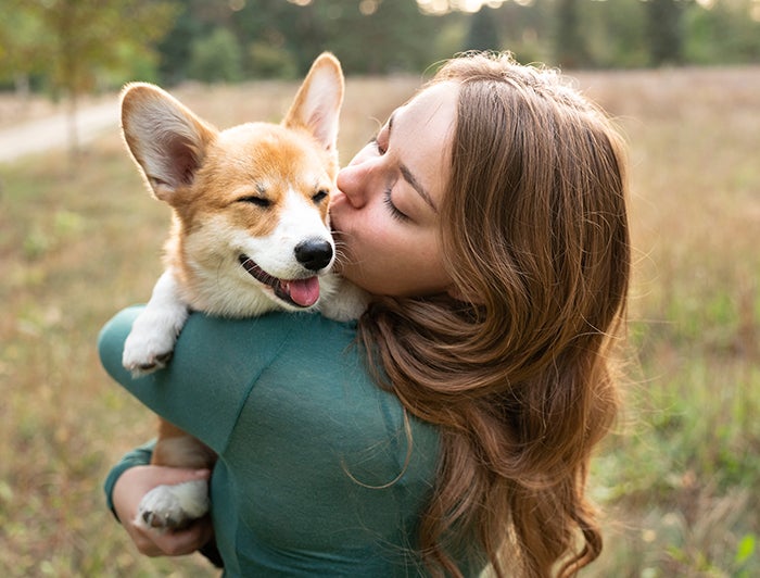 A young woman stands in a field holding and kissing a Corgi puppy