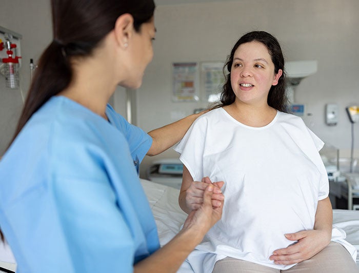MercyOne. Pregnant woman wearing hospital gown sits on edge of bed while holding the hand of a woman in scrubs