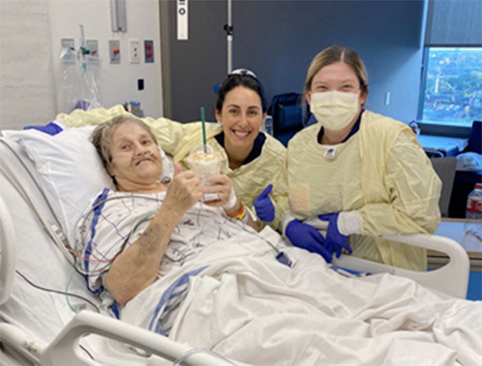 Pen medicine. Older female patient sits smiling in hospital bed holding a frappuccino while her caregivers stand beside her, smiling