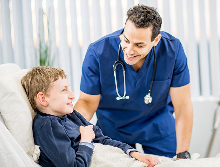 University of California Davis Health. Male physician wearing scrubs stands at a hospital bedside smiling down at the frail boy sitting up in the bed.