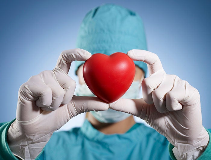 Tufts. Stock image shows a surgeon in scrubs holding a toy heart aloft, masking face