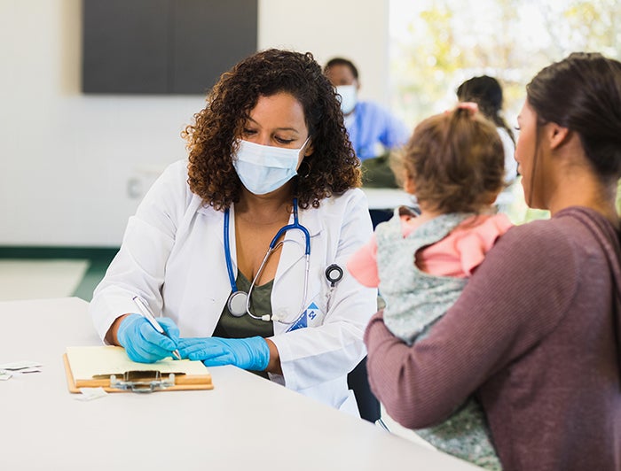 Providing Whole-Person Care by Addressing Patients’ Social and Economic Well-being: A female physician wearing medical mask sits filling out forms with a mother and child
