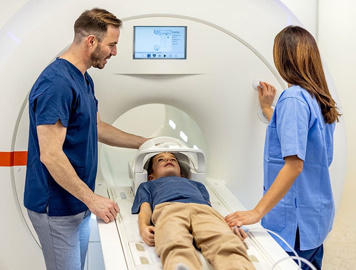 A man and a female health worker stand on either side of a boy lying in an MRI machine
