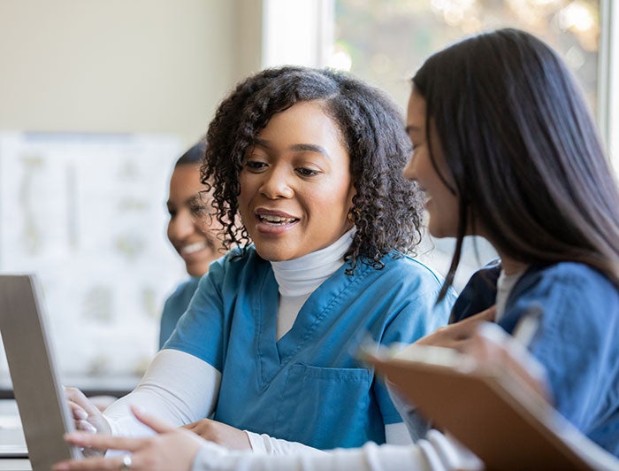 Two women wearing scrubs sit talking and gesturing towards an open laptop