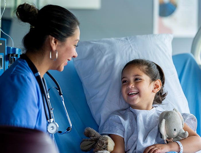 a little girl sits in a hospital bed smiling up at a female health worker