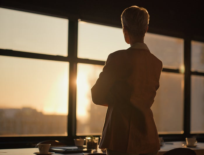 A woman stands staring out a window at sunset
