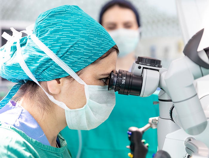 A female health worker wearing scrubs peers into a microscope in surgery