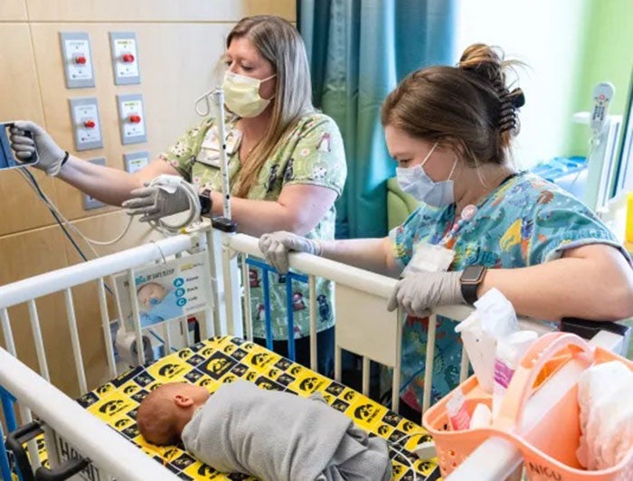 Two health workers attend to a swaddled infant in a hospital crib